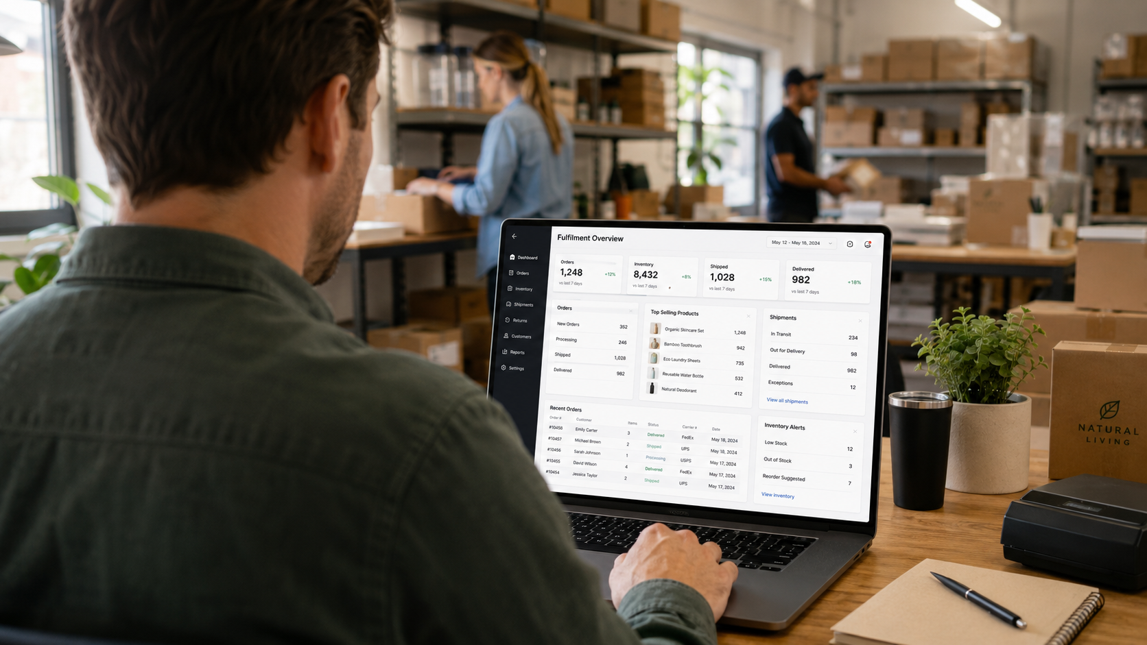 Person at a desk in a warehouse looking at a laptop with a 'Fulfillment Overview' dashboard on screen amidst shelves and boxes in the background.